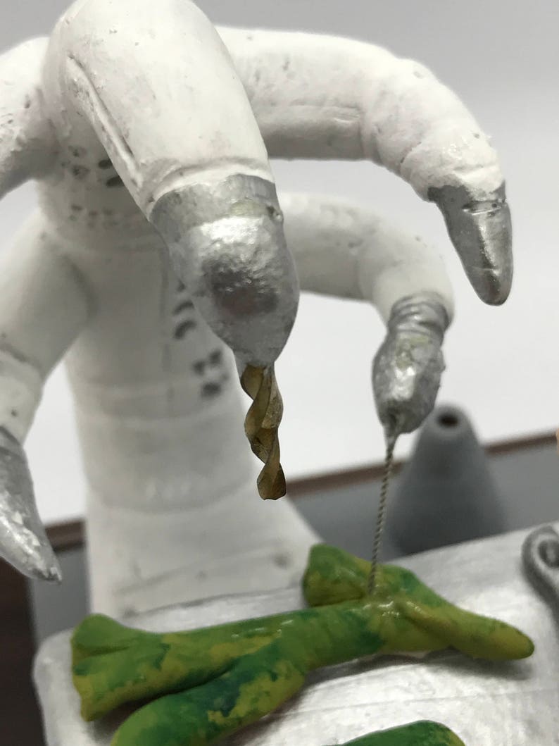 K&ouml;nnte beinhalten: Ein wei&szlig;er Roboterarm mit einer silbernen Spitze h&auml;lt einen Bohrer &uuml;ber einem gr&uuml;nen, strukturierten Objekt. Der Roboterarm ist &uuml;ber einem Tisch mit einer silbernen Oberfl&auml;che positioniert.