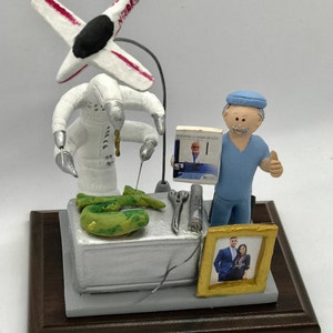 K&ouml;nnte beinhalten: Eine Figur eines Arztes in blauen OP-Kleidung mit erhobenem Daumen, der neben einem wei&szlig;en Roboter steht, der chirurgische Instrumente h&auml;lt. Der Roboter operiert an einem gr&uuml;nen Objekt. Der Arzt h&auml;lt ein Buch mit dem Titel "Building Your Health" mit einem Bild eines Mannes auf dem Cover. Im Hintergrund befindet sich ein gerahmtes Bild eines Paares.