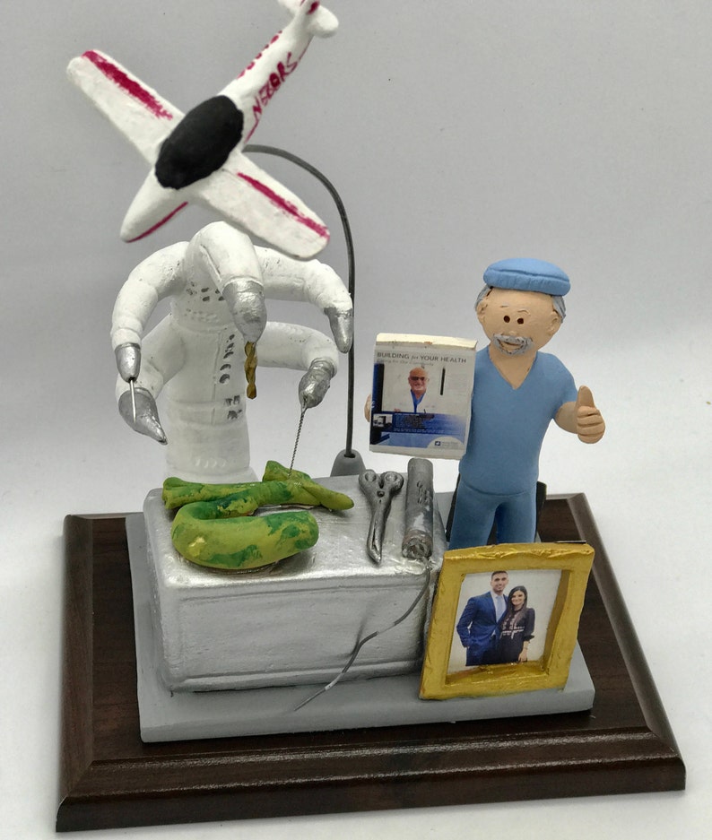 K&ouml;nnte beinhalten: Eine Figur eines Arztes in blauen OP-Kleidung mit erhobenem Daumen, der neben einem wei&szlig;en Roboter steht, der chirurgische Instrumente h&auml;lt. Der Roboter operiert an einem gr&uuml;nen Objekt. Der Arzt h&auml;lt ein Buch mit dem Titel "Building Your Health" mit einem Bild eines Mannes auf dem Cover. Im Hintergrund befindet sich ein gerahmtes Bild eines Paares.