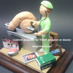 K&ouml;nnte beinhalten: Eine ma&szlig;geschneiderte, geformte Figur eines Arztes in gr&uuml;ner Sch&uuml;rze, der einen medizinischen Eingriff durchf&uuml;hrt. Die Figur h&auml;lt ein medizinisches Werkzeug und steht neben einem Tisch mit medizinischen Utensilien. Die Figur steht auf einem Holzsockel mit einer Plakette, auf der "custom sculpted for YOUR surgeon or doctor magicmud.com" steht.
