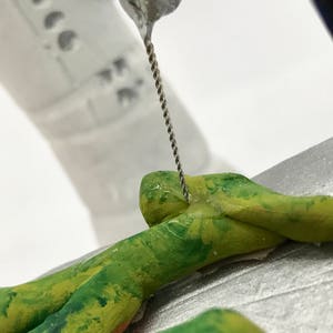 K&ouml;nnte beinhalten: Nahaufnahme einer gr&uuml;nen und gelben Tonfigur, die mit einem Metallwerkzeug modelliert wird. Das Werkzeug wird verwendet, um den Ton zu gl&auml;tten und eine klarere Form zu schaffen.