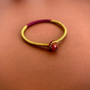 May include: A handmade bracelet featuring a woven design in gold and burgundy. The bracelet has a circular shape with a small, round, reddish-brown bead as a clasp. The bracelet is likely made of cord or thread.