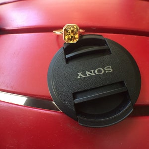 May include: A gold ring featuring a rectangular, faceted yellow gemstone in a bezel setting. The ring is resting on a black circular camera lens cap with the word SONY printed in white letters, providing a clear scale for the jewelry piece.