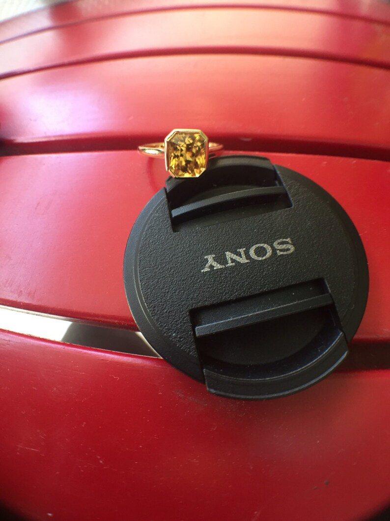 May include: A gold ring featuring a rectangular, faceted yellow gemstone in a bezel setting. The ring is resting on a black circular camera lens cap with the word SONY printed in white letters, providing a clear scale for the jewelry piece.