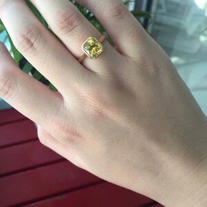May include: A gold ring featuring a faceted, square-cut yellow gemstone set in a smooth bezel setting. The band is a simple, polished gold metal. The stone displays a vibrant, translucent yellow hue with visible internal facets and light reflection.