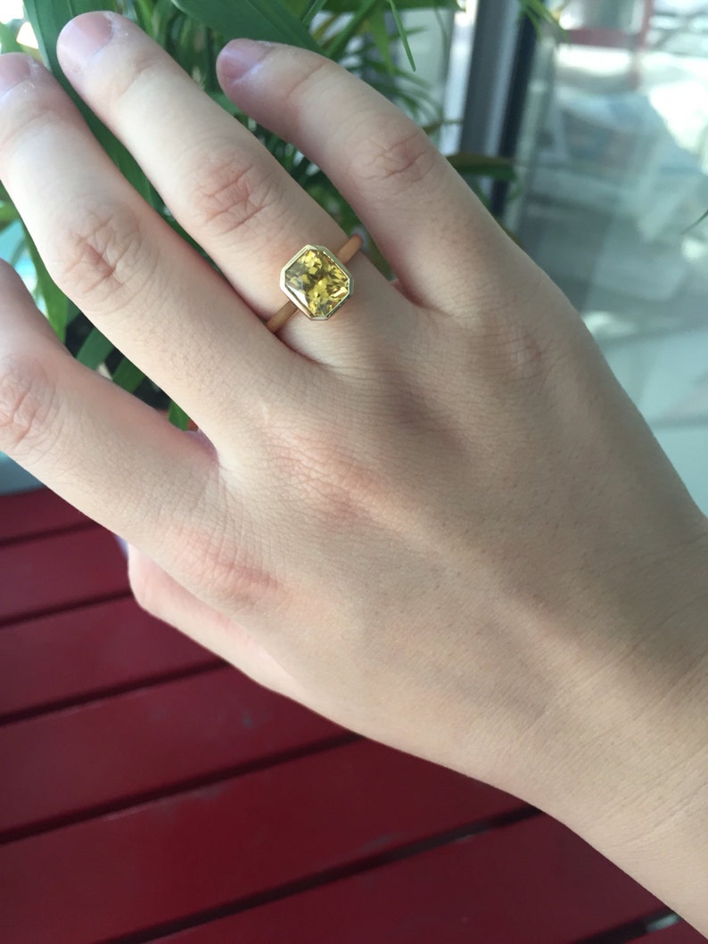 May include: A gold ring featuring a faceted, square-cut yellow gemstone set in a smooth bezel setting. The band is a simple, polished gold metal. The stone displays a vibrant, translucent yellow hue with visible internal facets and light reflection.