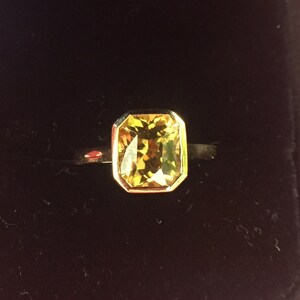 May include: A ring featuring a rectangular, faceted yellow gemstone set in a gold-toned bezel setting. The stone displays a warm, golden-yellow hue with visible internal facets that catch the light, mounted on a simple, smooth, dark-toned metal band.