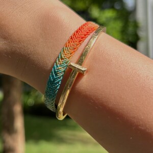 May include: Two bracelets on a wrist. One is a woven friendship bracelet with a gradient of colors from orange to teal. The other is a gold-colored metal bangle with a T-shaped accent.