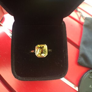 May include: A gold ring featuring a rectangular, faceted yellow-green gemstone set in a bezel setting. The ring is displayed inside a black velvet-lined jewelry box, highlighting the contrast between the metallic gold band and the gemstone.