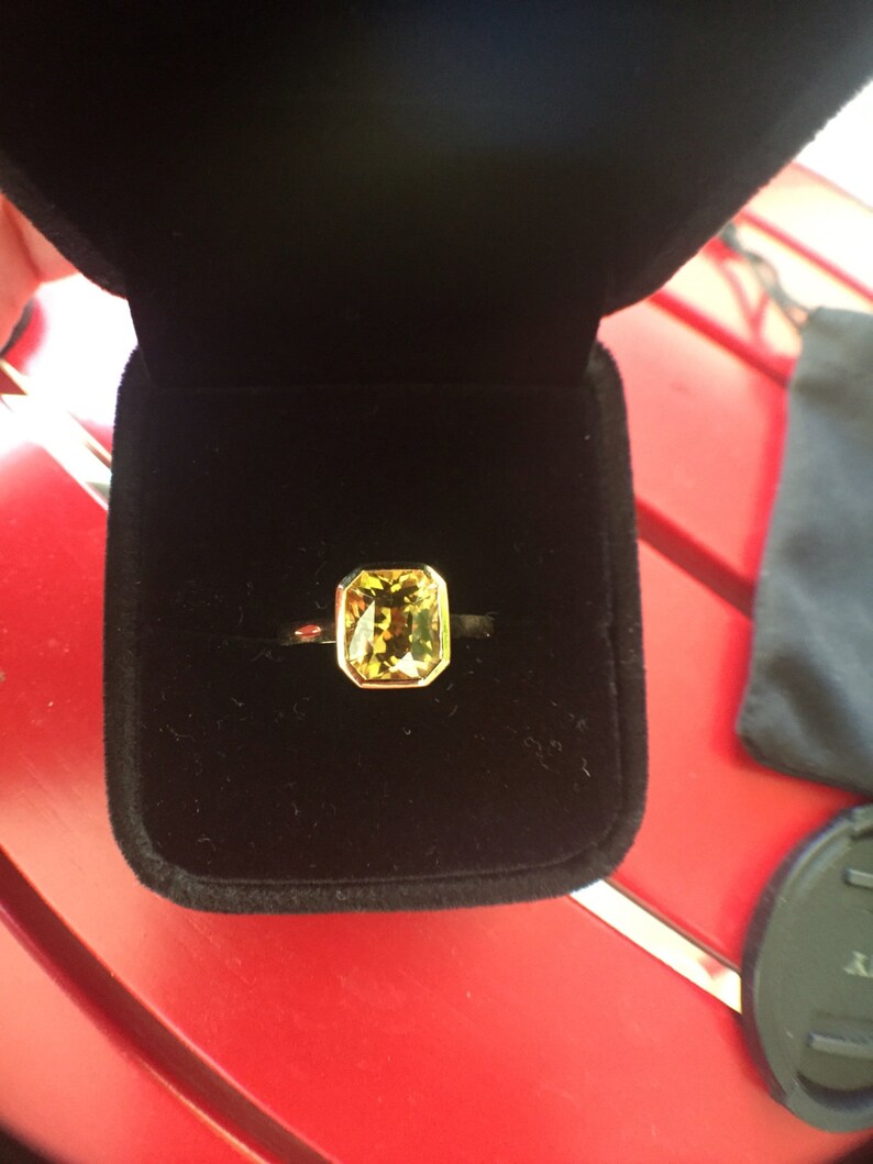 May include: A gold ring featuring a rectangular, faceted yellow-green gemstone set in a bezel setting. The ring is displayed inside a black velvet-lined jewelry box, highlighting the contrast between the metallic gold band and the gemstone.