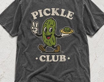 May include: A charcoal grey short-sleeved t-shirt featuring a vintage-style graphic of a green cartoon pickle character. The pickle is making a peace sign and holding a small plate. The text PICKLE CLUB is printed in a cream-coloured arched font.