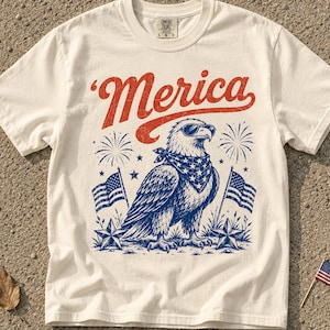 May include: An off-white short-sleeve t-shirt featuring a graphic of a bald eagle wearing sunglasses and a star-patterned bandana. The design includes the red text 'Merica', two American flags, stars, and fireworks in red and blue ink.