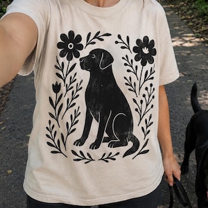 May include: Beige t-shirt with a black dog design framed by flowers. The dog is depicted sitting. The person is walking a black dog on a leash. The background shows a path through a green, tree-lined area.