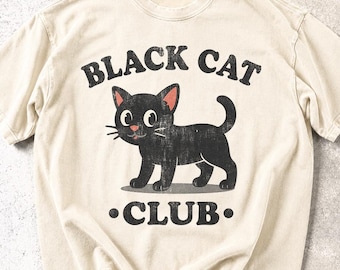 May include: A cream-colored short-sleeve t-shirt featuring a distressed black graphic of a cartoon cat. The text above the cat reads BLACK CAT and the text below reads CLUB. The design has a vintage, weathered appearance with a round neckline.