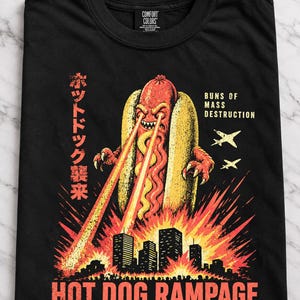 Puede incluir: Una camiseta negra con un gráfico de un monstruo hot dog gigante con ojos láser destruyendo una ciudad. El texto dice "BUNS OF MASS DESTRUCTION", "HOT DOG RAMPAGE", "MUSTARD-FUELED MAYHEM. YOUR INSURANCE WON'T COVER IT." y caracteres japoneses.