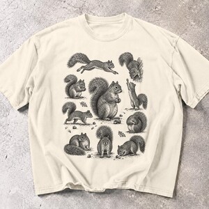 Può includere: Una t-shirt a maniche corte color crema con un'illustrazione dettagliata a inchiostro nero di vari scoiattoli in diverse pose, tra cui correre, sedersi e cercare cibo, distribuite sulla parte anteriore del capo.