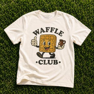 Puede incluir: Camiseta color crema con un personaje de gofre de dibujos animados haciendo un gesto de pulgar hacia arriba y sosteniendo una jarra peque&ntilde;a. El texto "WAFFLE CLUB" est&aacute; impreso arriba y abajo del personaje.