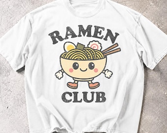 May include: A white short-sleeve t-shirt featuring a graphic of a smiling ramen bowl with arms and legs. The text RAMEN CLUB is printed in a distressed black font above and below the bowl, which contains noodles, an egg, and a fish cake.