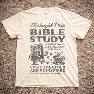May include: Cream-colored t-shirt with a distressed black graphic. The graphic reads "Midnight Drip BIBLE STUDY" with illustrations of a smiling Bible, coffee cup, and coffee beans. Text includes "Draw near to God" and "Come together and be inspired."