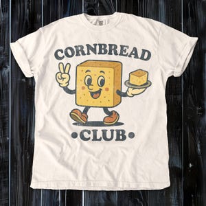 May include: Cream-colored t-shirt with a retro-style graphic. The design features a smiling, anthropomorphic cornbread character giving a peace sign and holding a slice of cornbread. The text "CORNBREAD CLUB" is arched above and below the character.