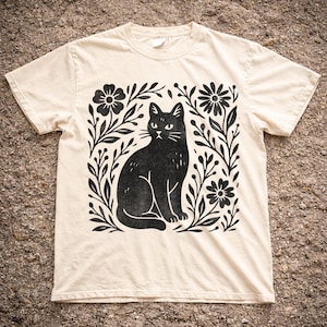 Könnte beinhalten: Cremefarbenes T-Shirt mit einer schwarzen Katzenillustration, umgeben von floralen und Blattmustern. Die Katze ist sitzend dargestellt, mit detaillierten Merkmalen. Das Design ist mittig auf dem Shirt platziert und erzeugt eine ansprechende Grafik.