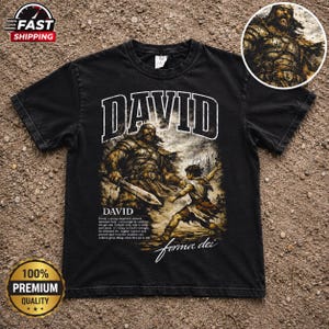 Puede incluir: Una camiseta negra de manga corta con un estampado gráfico de un guerrero y un joven, con el texto DAVID, un párrafo descriptivo y el texto en cursiva forma dei. Un recuadro circular muestra un primer plano del guerrero.