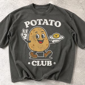 Könnte beinhalten: Ein anthrazitfarbenes Kurzarm-T-Shirt mit einer Grafik einer lächelnden Cartoon-Kartoffel. Die Kartoffel macht ein Friedenszeichen und hält einen Teller mit einem Stück Butter. Der Text POTATO CLUB ist in Weiß darüber und darunter gedruckt.