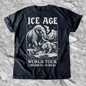Op de afbeelding: Zwart T-shirt met een gedetailleerde afbeelding van een wolharige mammoet en een sabeltandtijger op een ijzig landschap. De tekst "ICE AGE WORLD TOUR" staat erboven en "2.000.000 v.Chr. - 10.000 v.Chr." eronder.