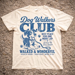 Könnte beinhalten: Cremefarbenes T-Shirt mit blauem Aufdruck "Dog Walkers Club". Das Design zeigt einen Cartoon-Hund an der Leine, Pfotenabdrücke und den Text "Walked & Wonderful".
