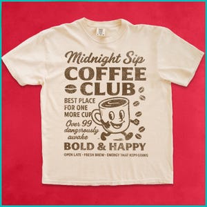 May include: Off-white t-shirt with a retro coffee-themed design. The graphic includes the text "Midnight Sip Coffee Club" and a cartoon coffee cup. Additional text reads "Best Place for One More Cup" and "Bold & Happy."