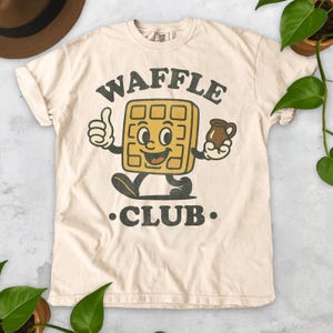 K&ouml;nnte beinhalten: Cremefarbenes T-Shirt mit einem Retro-Grafikdesign eines l&auml;chelnden Waffel-Charakters, der einen Daumen hoch zeigt und einen Sirupkrug h&auml;lt. Der Text "WAFFLE CLUB" ist in einer Vintage-Schriftart &uuml;ber und unter der Figur gedruckt.