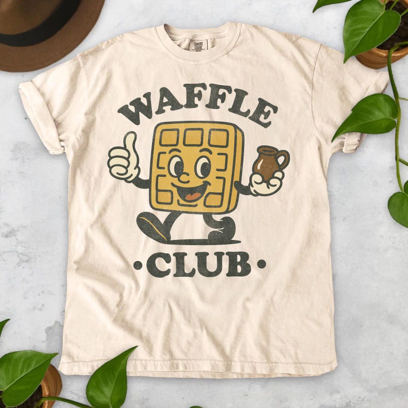 K&ouml;nnte beinhalten: Cremefarbenes T-Shirt mit einem Retro-Grafikdesign eines l&auml;chelnden Waffel-Charakters, der einen Daumen hoch zeigt und einen Sirupkrug h&auml;lt. Der Text "WAFFLE CLUB" ist in einer Vintage-Schriftart &uuml;ber und unter der Figur gedruckt.