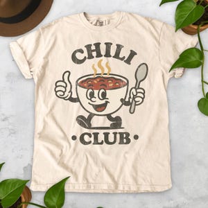 Op de afbeelding: Cr&egrave;mekleurig T-shirt met een vintage-stijl grafische afbeelding. Het ontwerp toont een lachende kom chili met armen en benen, die een lepel vasthoudt en een duim omhoog steekt. De tekst "CHILI CLUB" staat boven en onder de kom.