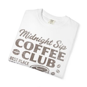 May include: White t-shirt featuring the text "Midnight Sip Coffee Club" in a textured brown font. The design includes coffee beans and a coffee cup graphic. The shirt is folded and displayed on a white background.