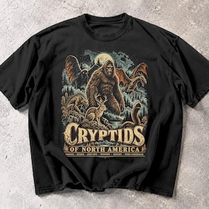 Puede incluir: Una camiseta negra de manga corta con un estampado gráfico de varios críptidos norteamericanos, incluyendo Bigfoot, Mothman y un jackalope. El texto dice "CRYPTIDS OF NORTH AMERICA" con una lista de nombres debajo de la ilustración.
