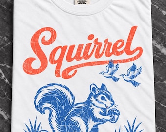 May include: A white t-shirt featuring the word Squirrel in a distressed orange script font. Below the text is a blue graphic of a squirrel holding an acorn, surrounded by small mushrooms, acorns, and two flying birds, all with a cracked texture.