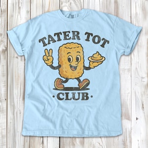 May include: Light blue t-shirt with the words "TATER TOT CLUB" in dark brown lettering. The shirt features a cartoon tater tot character with arms and legs, holding a plate of sauce and giving a peace sign.