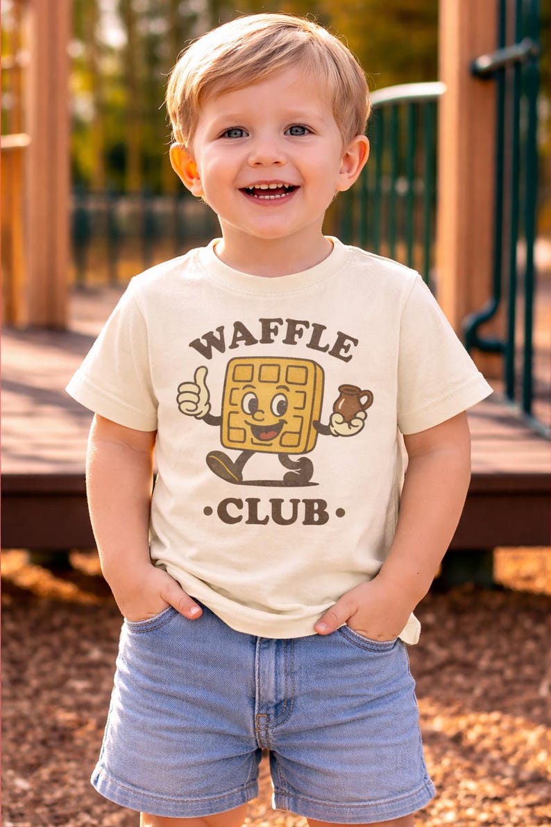 Puede incluir: Un ni&ntilde;o peque&ntilde;o con una camiseta color crema con un gr&aacute;fico de gofres y las palabras "WAFFLE CLUB". El personaje del gofre sonr&iacute;e y levanta el pulgar. El ni&ntilde;o tambi&eacute;n lleva pantalones cortos vaqueros azules.