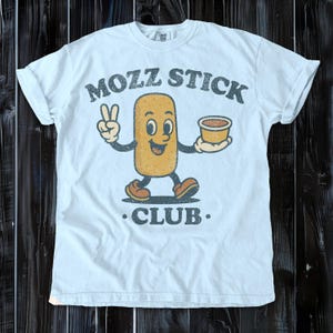 May include: Light blue t-shirt featuring a cartoon mozzarella stick character with a peace sign and a cup of dipping sauce. The text "MOZZ STICK CLUB" is printed above and below the character in a distressed font.