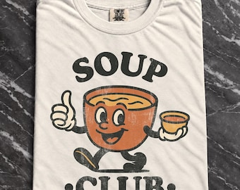 May include: A cream-coloured t-shirt featuring a graphic of a cartoon soup bowl with a face, arms, and legs. The bowl gives a thumbs up and holds a smaller cup. The text SOUP CLUB is printed in black above and below the graphic. The shirt has a Comfort Colors label.