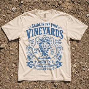 May include: Cream-colored t-shirt with a blue graphic design. The design features the words "Abide in the Vine Vineyards" and a cartoon grape character. Additional text includes "John 15:5" and "Apart from Him we can do nothing."