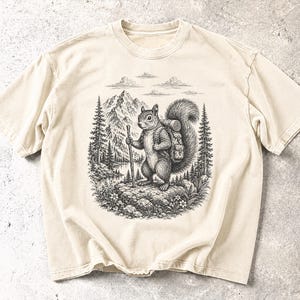 May include: A cream-coloured short-sleeve t-shirt featuring a detailed black ink illustration of a squirrel wearing a backpack and holding a walking stick. The scene includes a mountain range, pine trees, a lake, and clouds in the background.