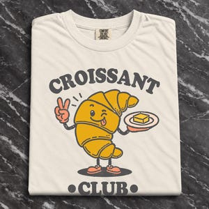 Könnte beinhalten: Ein cremefarbenes T-Shirt mit einer Grafik eines Cartoon-Croissants mit Gesicht, das zwinkert und die Zunge herausstreckt. Das Croissant hält einen Teller mit einem Stück Butter und zeigt ein Friedenszeichen. Der Text lautet CROISSANT CLUB.