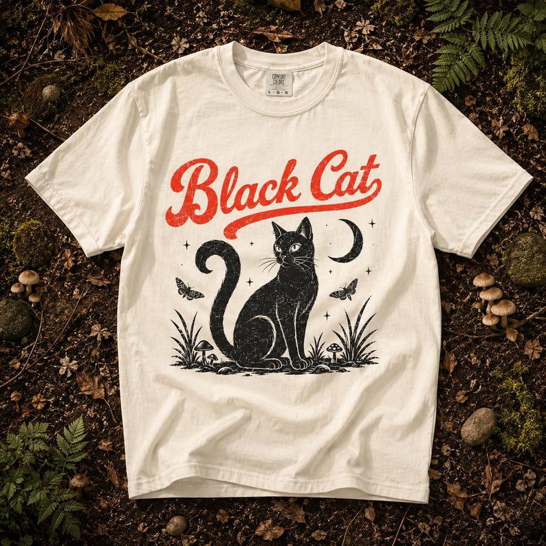 K&ouml;nnte beinhalten: Ein cremefarbenes T-Shirt mit kurzen &Auml;rmeln und einer Grafik eines schwarzen Katers, der zwischen Pilzen und Gras sitzt. &Uuml;ber dem Kater steht der Text Black Cat in einer abgenutzten roten Schrift. Das Design enth&auml;lt zudem einen Mond, Sterne und Motten.