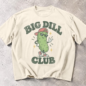 Puede incluir: Una camiseta de manga corta color crema con un gráfico de un pepinillo verde que lleva una gorra de béisbol roja, gafas de sol negras y zapatillas rojas. El pepinillo señala con una mano. El texto BIG DILL CLUB está impreso en verde oscuro.