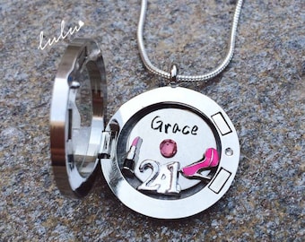 Puede incluir: Collar con dije de plata en forma de medallón con una piedra rosa y la palabra "Grace" grabada en la parte delantera. El medallón está abierto para revelar un dije de zapato rosa y el número "21" en el interior.