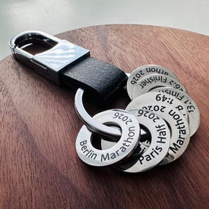 Op de afbeelding: Een sleutelhanger met een zwarte leren band en zilverkleurige metalen hardware. Bevestigd zijn meerdere ronde metalen tags met de inscripties "Berlin Marathon 2026", "Paris Half 2026" en andere racedetails.