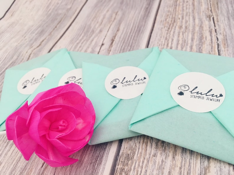 May include: Four light blue gift boxes with white circles and the text "lulula stamped jewellery" on each box. A pink flower is in the foreground.