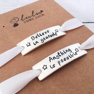 May include: Two silver bracelets with white ribbon ties. The bracelets are stamped with the words "Believe in yourself" and "Anything is possible" with a star on each.