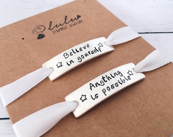 May include: Two silver bracelets with white ribbon ties. The bracelets are stamped with the words "Believe in yourself" and "Anything is possible" with a star on each.